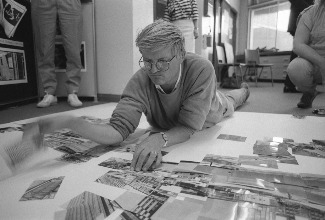 A black and white photography of David Hockney lying on the floor looking at images