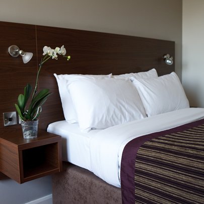 Double bed with white sheets, brown bedspread and flowers on the nightstand