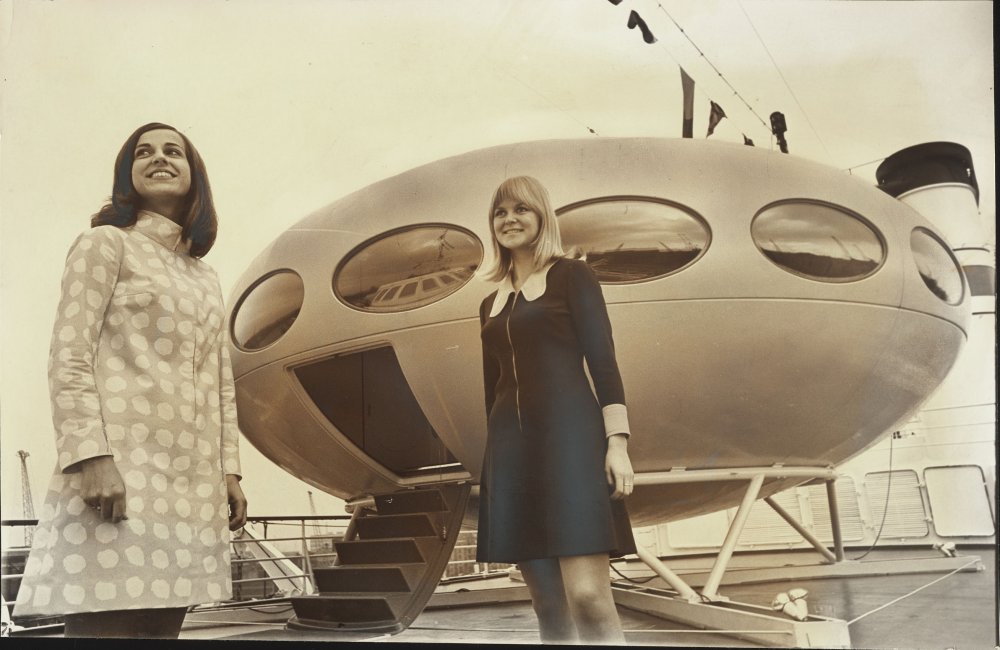 Two women standing in front of a 'Futuro House', a circular spaceship-esque pod with oval windows