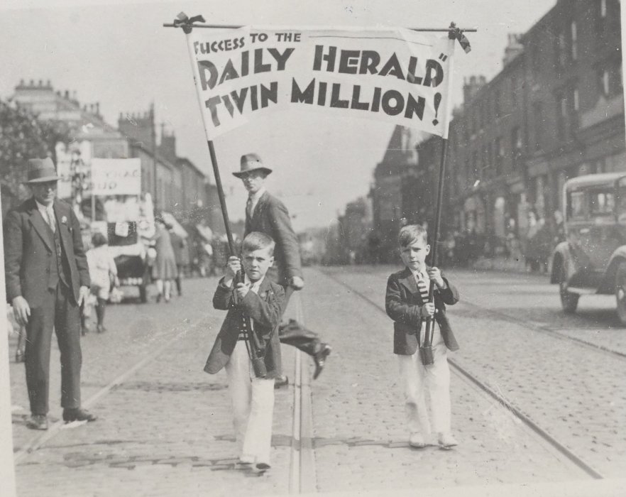 Two boys - twins in identical outfits - on an urban street, carrying a banner reading 'Success to the Daily Herald twin million!'