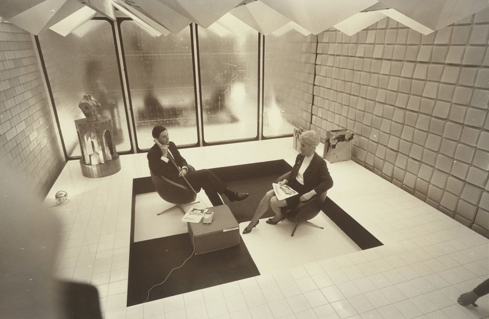 A man and woman sitting in a minimalist room with sunken central area and glass walls