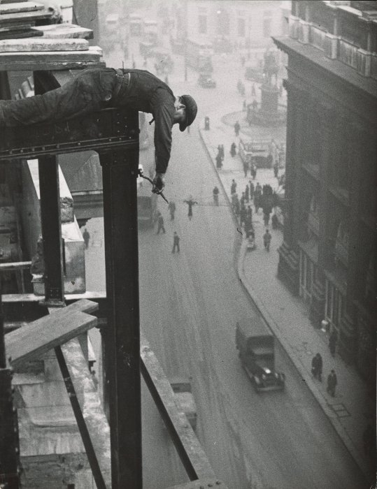 A steel erector leans over a building frame to tighten bolts, while pedestrians pass below