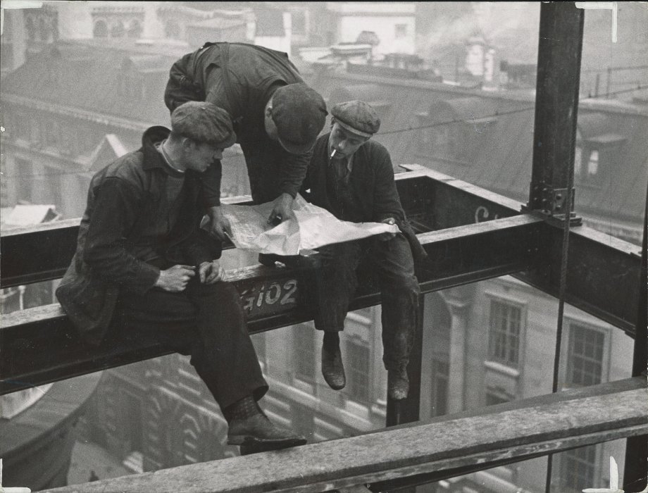 Three steel erectors examine plans while perched on a steel girder