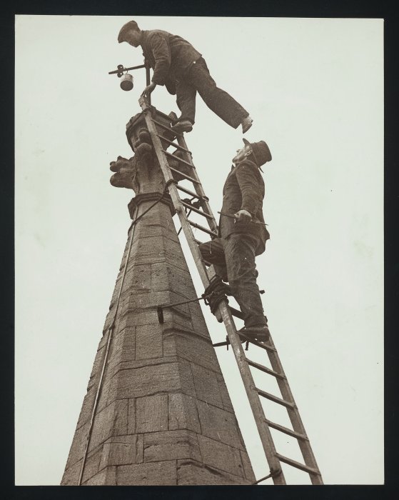 Two steeplejacks using a ladder affixed to the roof of a church to replace the weathercock