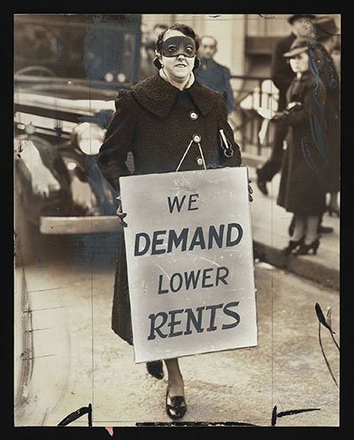 Masked demonstrator holding ‘We demand lower rents’ placard
