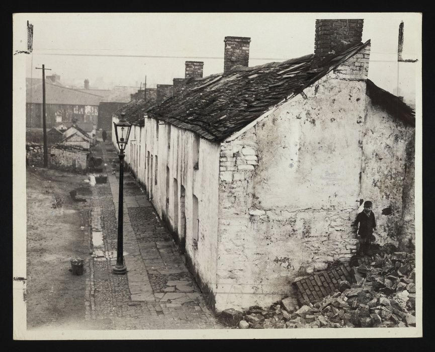 Young boy standing on a pile of rubble outside a row of miner’s cottages
