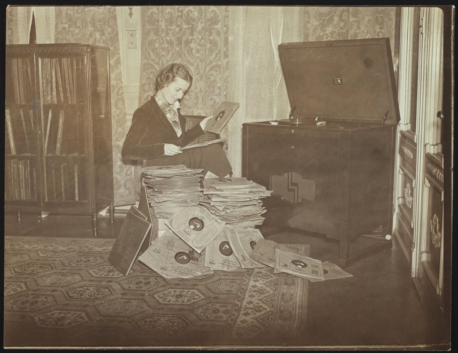A woman selects records to play on the large gramophone player in the corner of the room