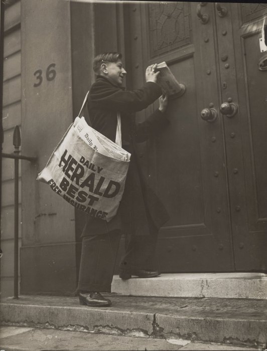 A young man, carrying a large bag of copies of the Daily Herald, pushes an issue of the newspaper through a letterbox