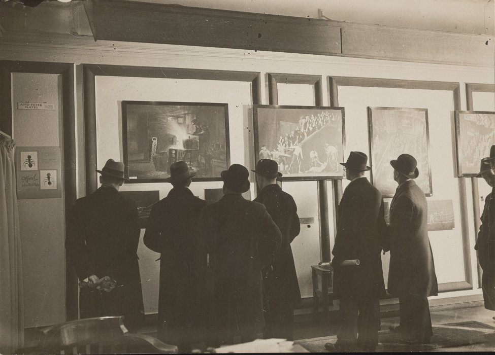 A crowd of men in coats of hats looking at photographs in a gallery