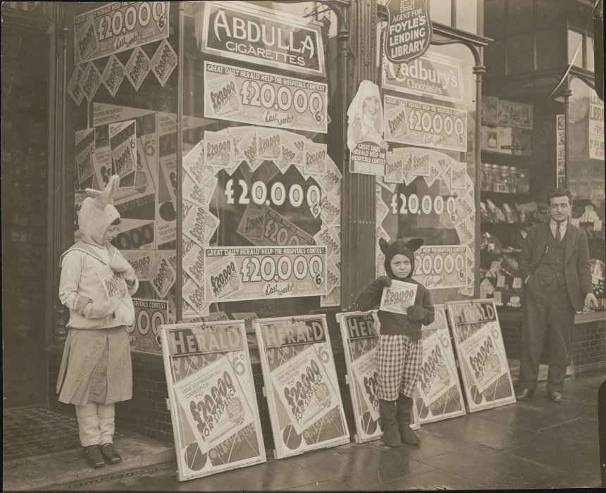 Two children in costumes standing outside a shop to promote a competition supporting local hospitals, with signs advertising £20,000 prize