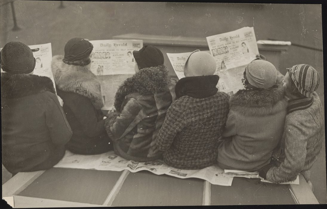A group of workers (shown from the back) reading the front page of the Daily Herald, announcing the results of a competition with a £25,000 prize