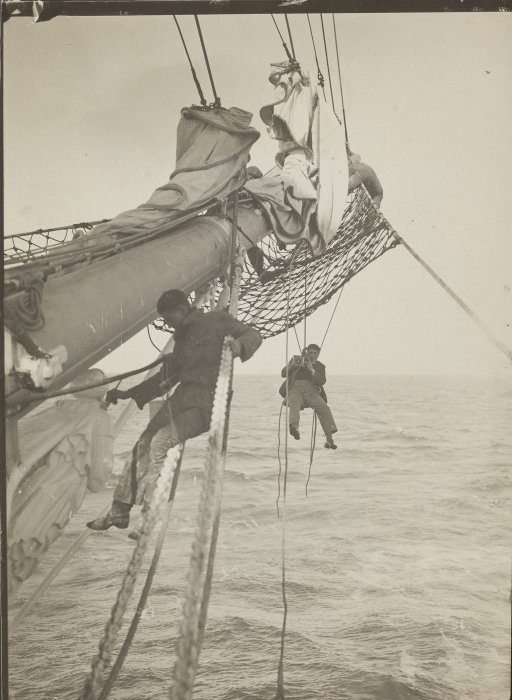 A man using a bosun’s chair, attached to a mast with ropes, to photograph a ship